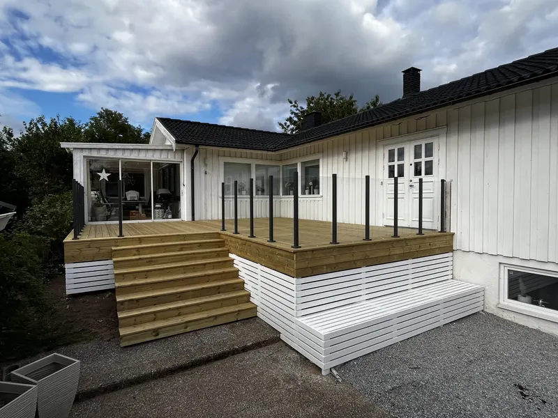 Altan i trä med glasräcke, svarta stolpar och bred trappa vid ett vitt hus